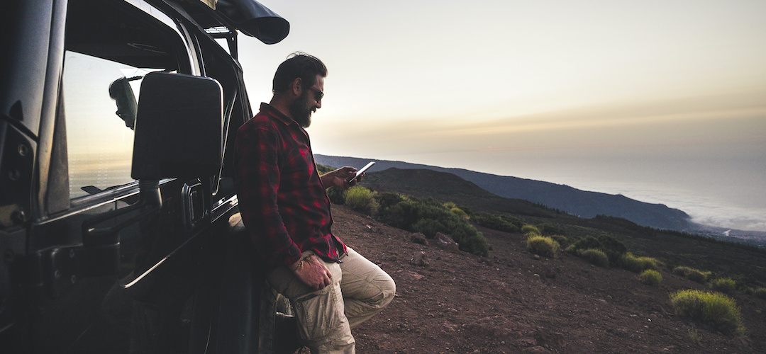 Adventure explorer traveler man use cellular phone with internet connection in wild mountain place during travel excursion with off road black car and tent on the roof - free people concept